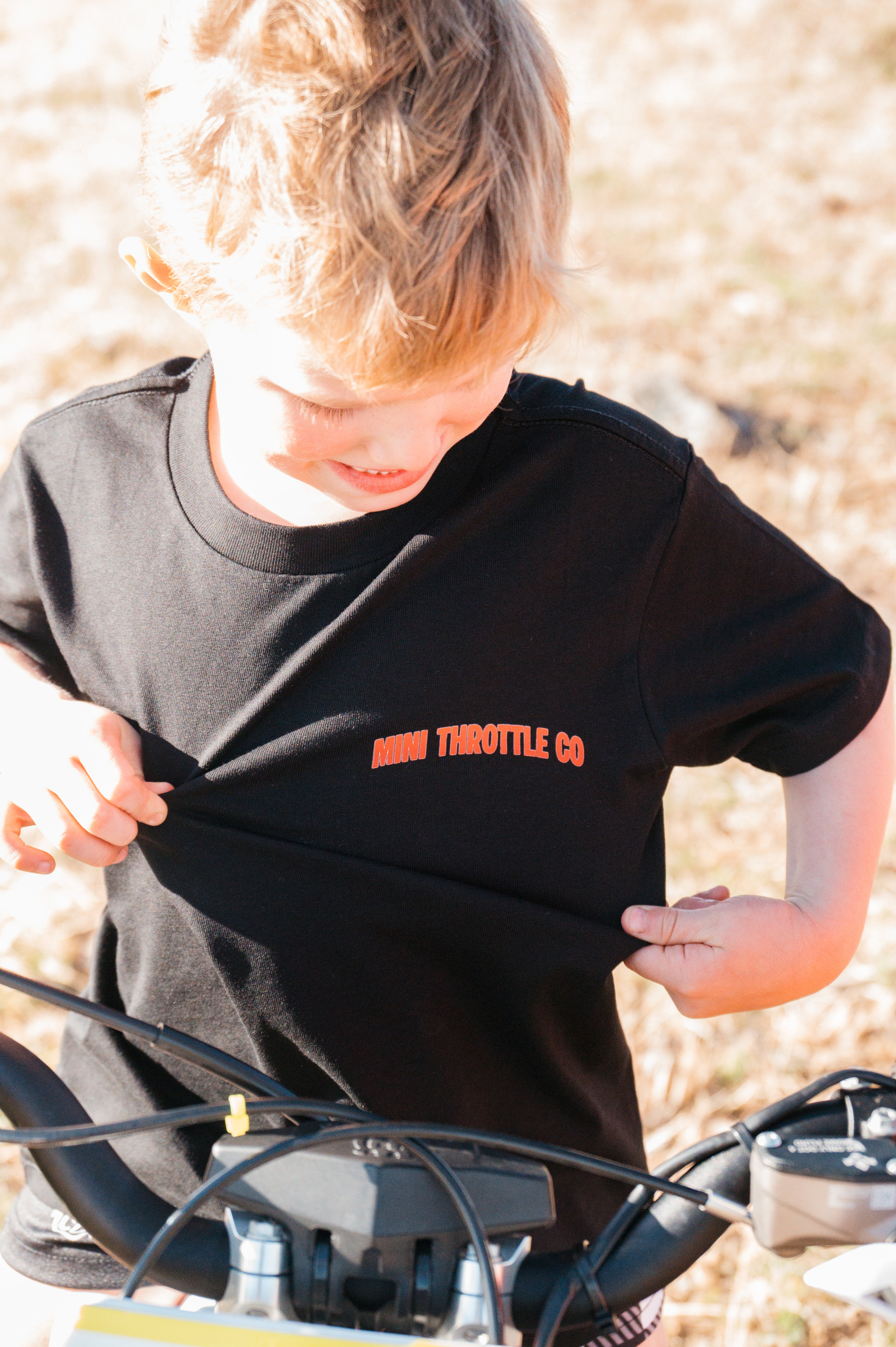 Child wearing a black t-shirt with 'Mini Throttle Co' text, standing outdoors.