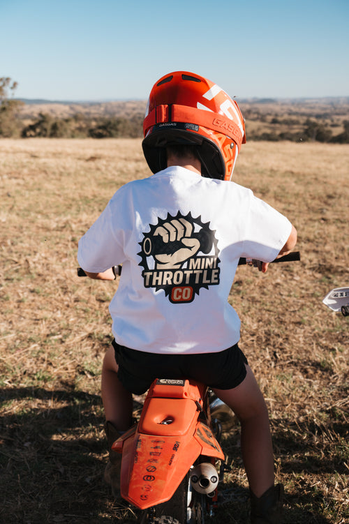 Child on an orange dirt bike wearing a helmet and a white shirt with a logo, standing in a dry field.