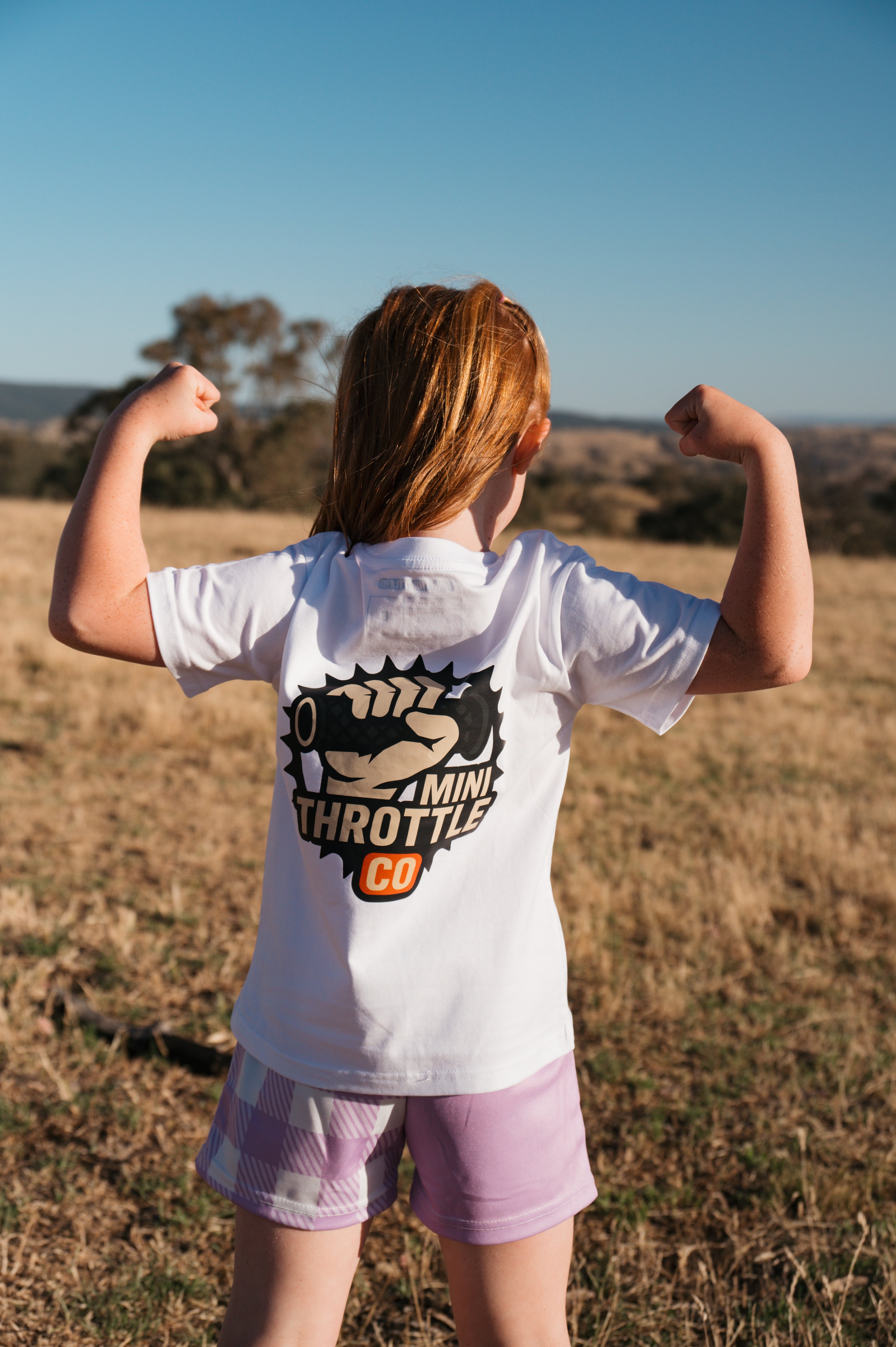 Young girl wearing a white t-shirt with a logo, flexing muscles in an open field.