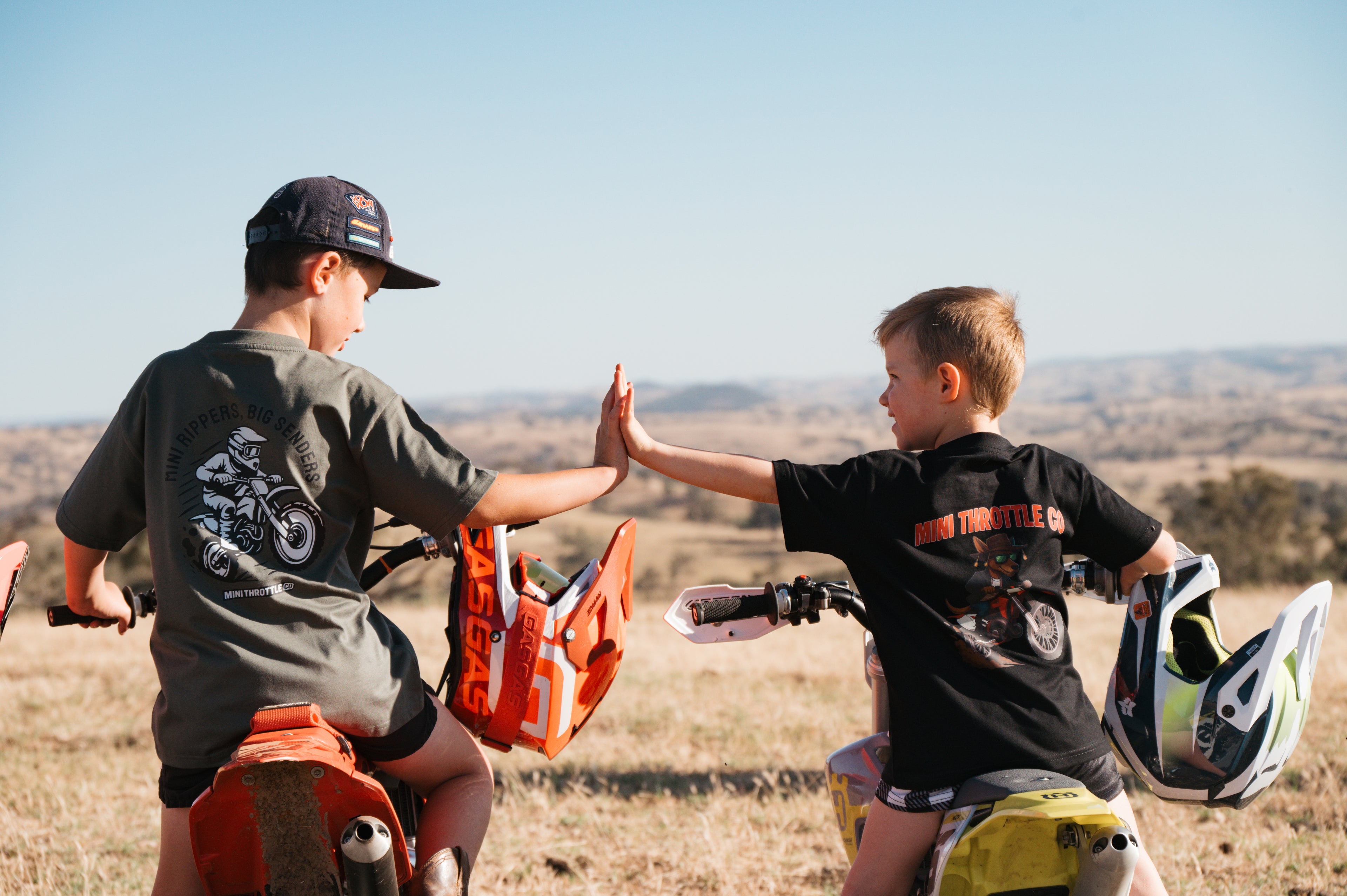 Two young boys on dirt bikes high-fiving each other in an open field.